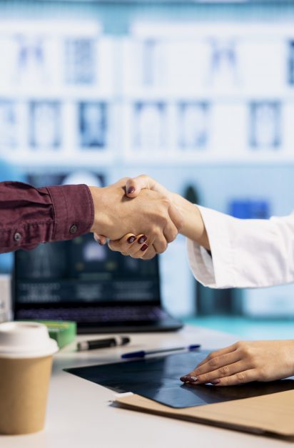 Indian general practitioner greeting her male patient at a medical examination, sharing a handshake and discussing about diagnostic reports. Woman doctor helping man to treat an illness. Close up.